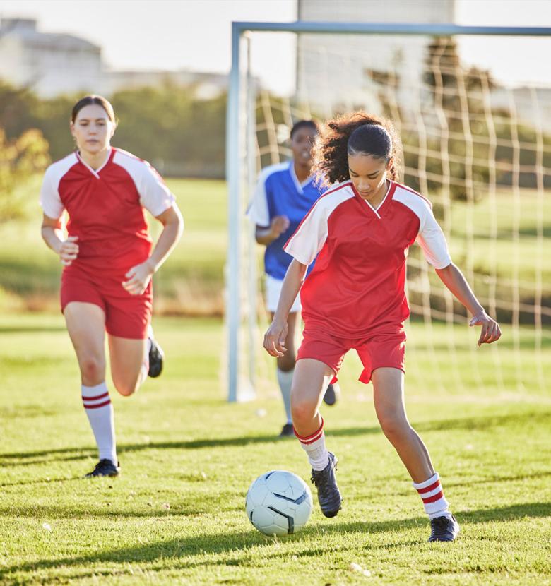 Girls playing soccer on a green field, wearing red and white uniforms.
